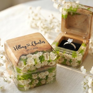 May include: Two square wooden ring boxes with floral detailing. One box is open, displaying a diamond ring on a black velvet base. The other box is closed, with the names "Hillary & Charles" and the date "09.17.2022" engraved on the lid.