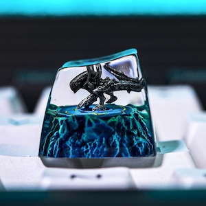 May include: A custom keyboard keycap with a detailed, dark-coloured alien figure set against a blue, textured background. The keycap is transparent, encased in clear resin, and sits on a white keyboard.