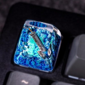 May include: A blue and green resin keycap with a silver sword design. The keycap is on a black keyboard.