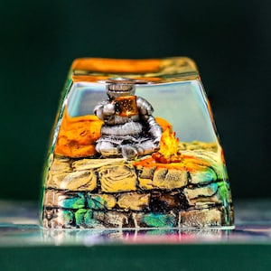 May include: A clear resin keycap with a miniature scene inside. The scene features a small figure sitting on a stone structure with a fire in front of them. The background is a green and brown landscape.
