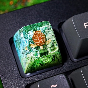 Puede incluir: Un tapón de tecla de resina verde y marrón con un diseño de tortuga. El tapón de tecla está en un teclado negro.