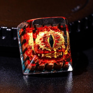 May include: A red and gold eye keycap with a black pupil, encased in clear resin.