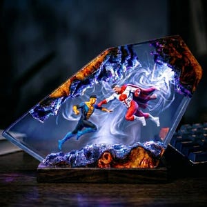 May include: A rectangular resin lamp featuring a dynamic scene of two figures in mid-air combat. One figure wears a blue and yellow suit, while the other wears a red and white suit with a cape. The resin contains swirling white smoke effects.