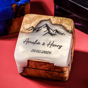 May include: Square wooden box featuring a mountain design, the names "Amelia & Henry", and the date "20.02.2025". The box has a white and brown colour scheme with a glossy finish. Two other boxes are visible in the background.