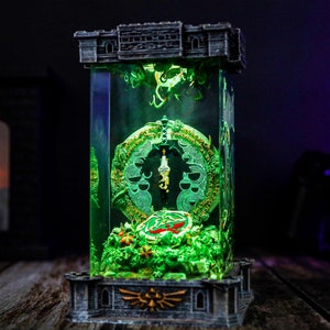 Puede incluir: Una escultura rectangular iluminada con un diseño verde brillante y una parte superior e inferior en forma de castillo. La escultura tiene un diseño detallado y ornamentado con un elemento circular central y una pequeña figura. La base tiene un emblema dorado.