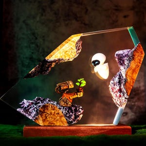 Könnte beinhalten: Dekorative Lampe mit Figuren aus dem Animationsfilm Wall-E. Die Lampe hat einen Holzsockel und eine klare, abgewinkelte Oberseite mit eingebetteten felsartigen Formationen. Die Figuren sind beleuchtet und erzeugen ein warmes Leuchten.