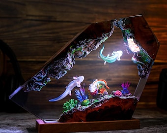 May include: A resin sculpture of a cave with three axolotls, a type of salamander, swimming inside. The cave is lit from within, casting a warm glow on the axolotls and the surrounding plants. The sculpture is mounted on a wooden base.