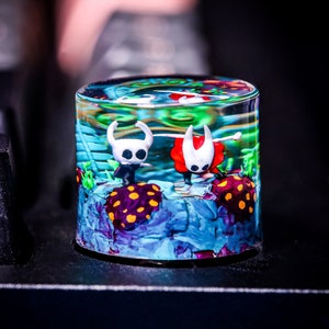 Può includere: Una cupola di vetro trasparente riempita di resina blu, verde e viola, con due piccole figurine bianche di personaggi del videogioco Hollow Knight. Le figurine sono posizionate come se stessero nuotando nella resina.