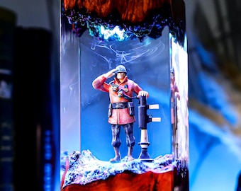 以下が含まれることがあります： 敬礼するミニチュア兵士を特徴とする長方形の樹脂アート作品。兵士は赤い制服を着て、岩の土台の上に立っています。樹脂ブロックは、暗いオレンジと青の色合いで、発光効果があります。