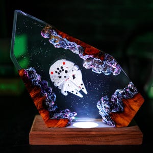 Könnte beinhalten: Ein einzigartiges Kunstwerk aus Harz mit einem weißen Millennium Falcon-Modell vor einem Sternenhimmel mit farbenfrohen Felsformationen. Das Kunstwerk ist in klarem Harz eingeschlossen und auf einem Holzsockel montiert.