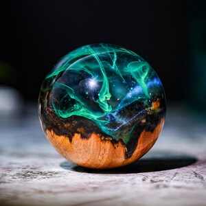 May include: A round, wooden sphere with a clear resin coating. The resin contains swirls of green and blue, resembling a galaxy. The sphere is approximately 2 inches in diameter.