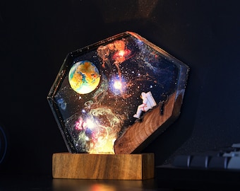 Könnte beinhalten: Eine sechseckige Harzlampe mit einer Galaxie-Szene, die einen Planeten, Sterne und eine kleine Astronautenfigur zeigt. Die Lampe ist beleuchtet und strahlt ein warmes Licht aus. Sie steht auf einem Holzsockel.