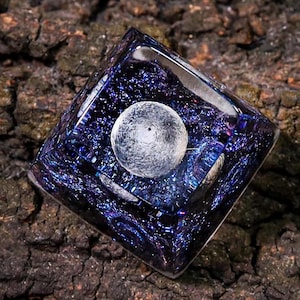 Peut inclure: Un capuchon de touche carré en résine transparente avec un design de type galaxie. Le capuchon de touche présente une lune argentée avec un centre sombre, entourée d'une nébuleuse bleue et violette tourbillonnante.