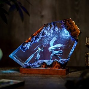 May include: A rectangular resin art piece featuring a battle scene with two figures, illuminated by blue light. The artwork is encased in clear resin with a wooden base and accents. The edges have a brown, textured appearance.