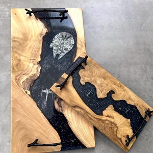 May include: Two rectangular serving trays made of natural light-coloured wood with a central river of dark blue resin embedded with white star-like flecks. The resin features miniature starship models and black metal handles shaped like tree branches.