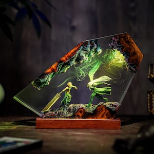 May include: A resin art piece featuring two figures in a battle scene, encased in clear resin. The figures are green and gold, set against a backdrop of green and orange rock formations. The base is made of wood.