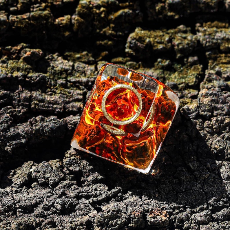 May include: A clear resin dice with a gold ring embedded inside. The dice is filled with orange and red flakes and is sitting on a dark brown textured surface.