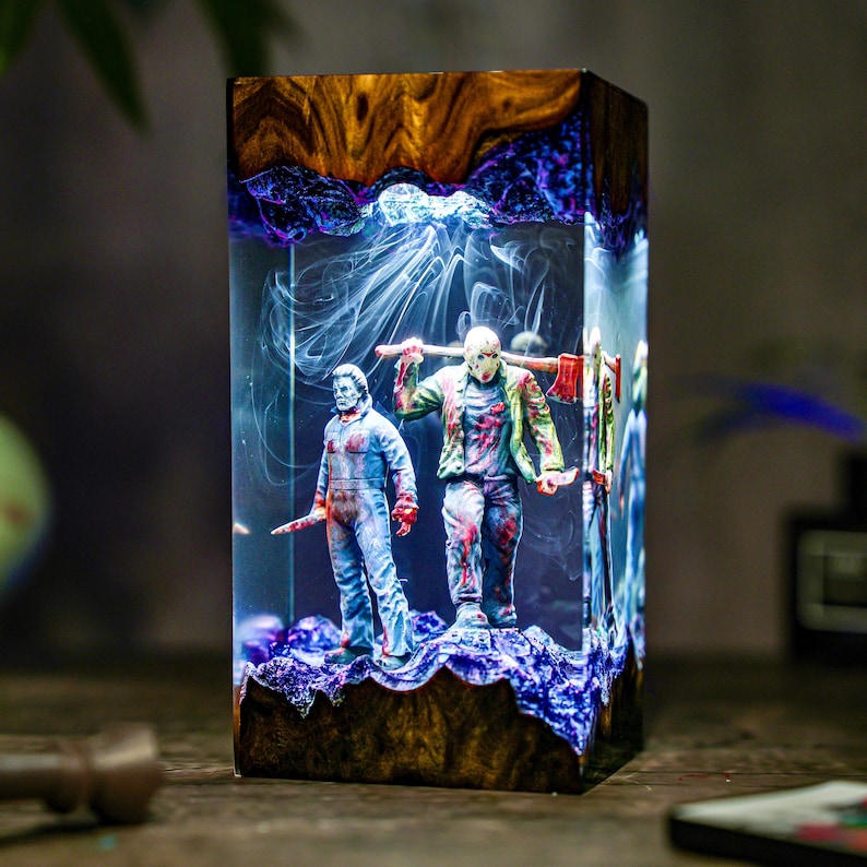 May include: A rectangular resin art piece featuring two horror movie figures. One figure wears a hockey mask and carries an axe, the other holds a knife. The piece has a wooden base and a glowing blue and white interior.