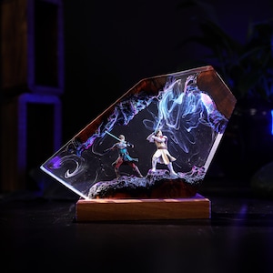 May include: A resin sculpture featuring two figures with lightsabers in a battle scene. The figures are set within a clear, angled resin block with a wooden base. The scene is illuminated with blue and purple hues.