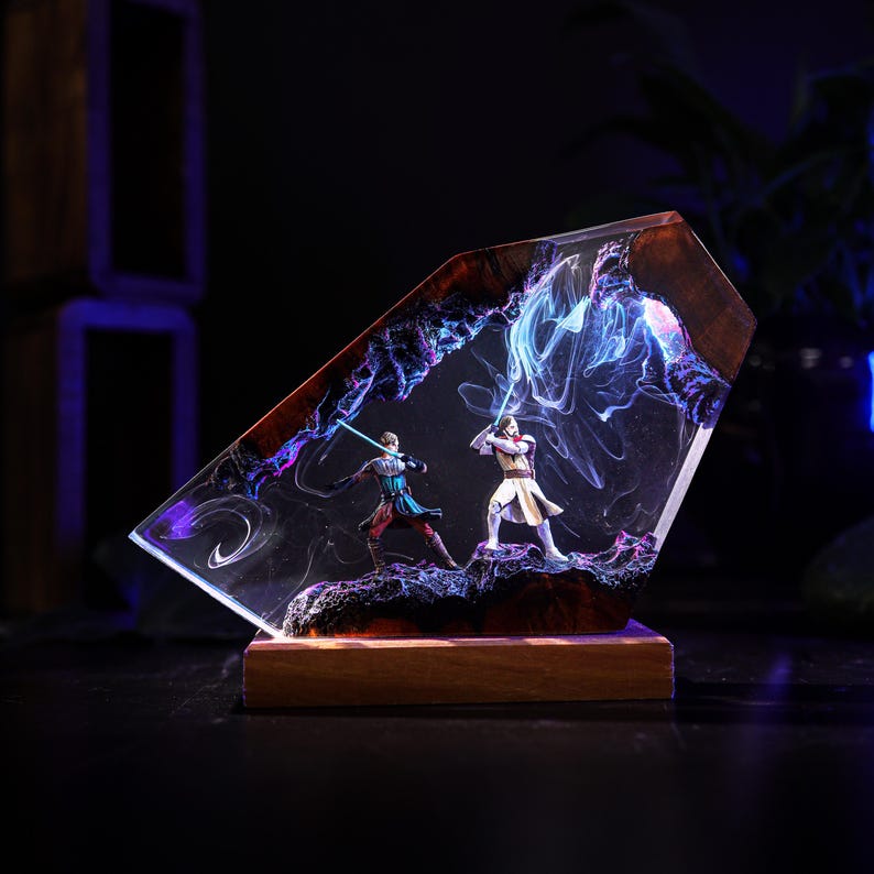 May include: A resin sculpture featuring two figures with lightsabers in a battle scene. The figures are set within a clear, angled resin block with a wooden base. The scene is illuminated with blue and purple hues.