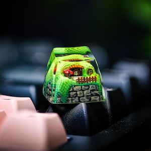 May include: A green and brown resin keycap with a miniature house and a small garden inside. The keycap is sitting on a black keyboard.