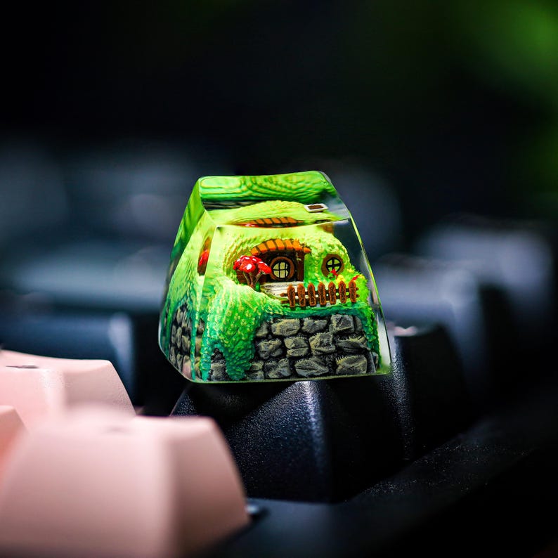 May include: A green and brown resin keycap with a miniature house and a small garden inside. The keycap is sitting on a black keyboard.