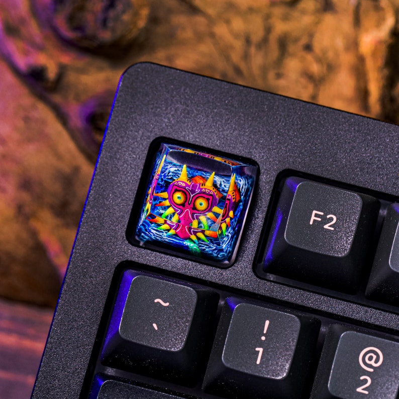 May include: A black keyboard with a custom keycap featuring a colorful illustration of Majora's Mask from the Legend of Zelda video game series. The keycap is a square shape and is made of clear plastic with a 3D design.