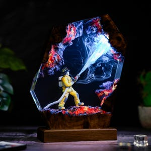 May include: A decorative lamp featuring a miniature firefighter in yellow gear standing on a dark, textured base. The scene is encased in clear resin with wood accents, illuminated from within to create a glowing effect of water spraying.