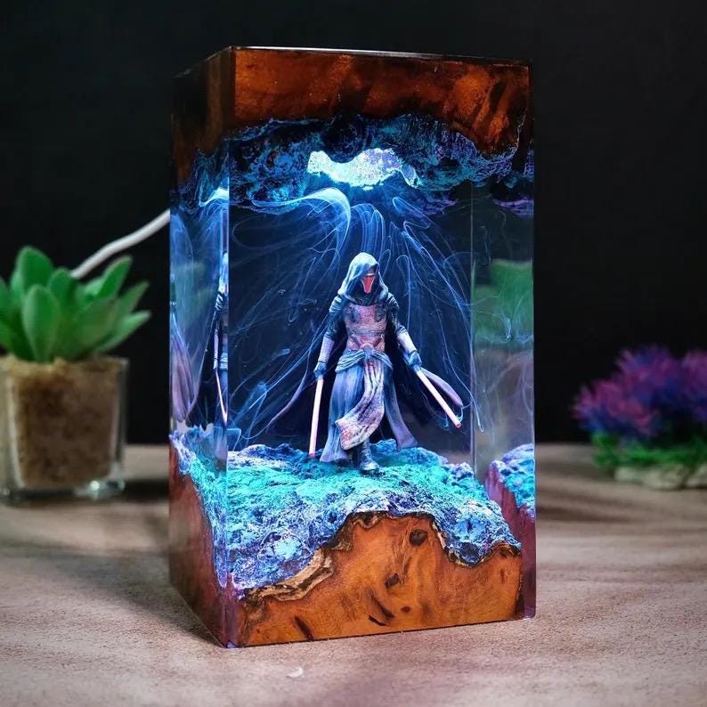May include: A rectangular resin and wood lamp featuring a hooded figure with two glowing light sabers standing on a textured blue base. The interior is filled with swirling blue smoke effects, encased in clear resin with natural wood top and bottom.