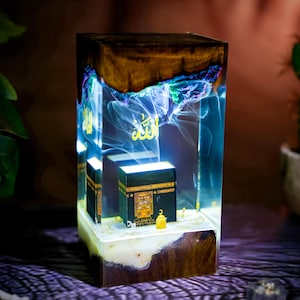 May include: A rectangular resin lamp featuring a dark wood base and top, with a clear center containing a miniature black Kaaba model and a small gold lantern. The Arabic word Allah is illuminated in gold, surrounded by swirling blue and white smoke.