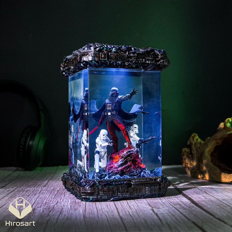 K&ouml;nnte beinhalten: Eine gl&auml;serne Vitrine mit schwarzem Sockel und durchsichtiger Oberseite. In der Vitrine steht eine dunkle Figur von Darth Vader mit einem roten Lichtschwert. Zwei wei&szlig;e Sturmtruppen stehen hinter ihm. Der Sockel der Vitrine ist mit einem rot-schwarzen lavaartigen Material verziert.