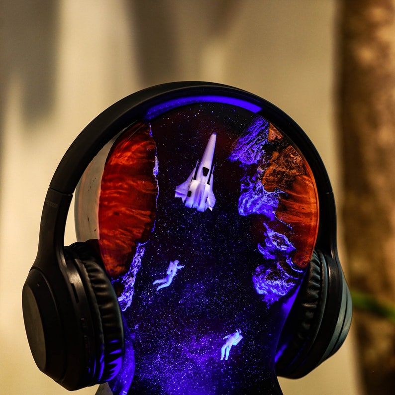 May include: Black headphones with a resin base that features a galaxy-like design with a white spaceship and astronauts. The resin base is illuminated with blue and purple lights.