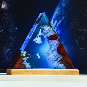 May include: A triangular resin sculpture with a blue and white astronaut and spaceship inside. The resin is clear and has a wood-like texture on the outside. The sculpture is on a wooden base.