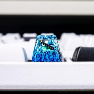May include: A blue and green resin keycap with a clear top and a black metal accent. The keycap is sitting on a white keyboard.