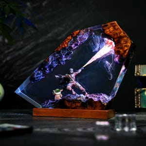 May include: A decorative resin and wood lamp featuring a miniature armored figure in a dynamic pose firing a bright orange light beam, accompanied by a small green creature. The scene is encased in a translucent, angular block with dark wood.