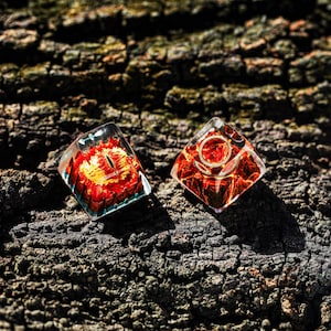 May include: Two clear resin dice with red and orange swirls. The dice are shaped like a pyramid with a rounded top. The dice are sitting on a dark brown tree bark surface.