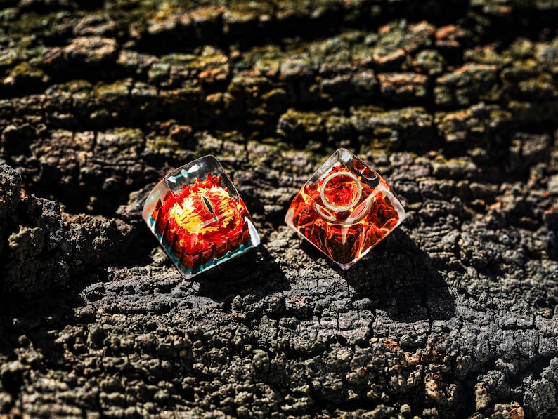 May include: Two clear resin dice with red and orange swirls. The dice are shaped like a pyramid with a rounded top. The dice are sitting on a dark brown tree bark surface.