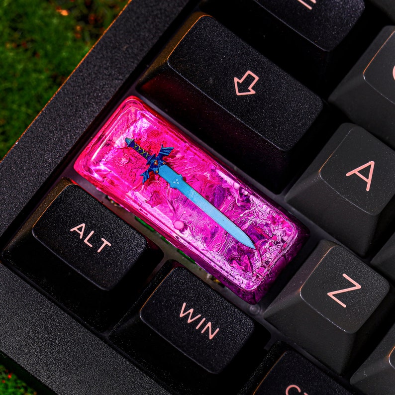 May include: A pink and blue resin keycap with a blue sword design. The keycap is on a black keyboard with the keys labeled "ALT", "WIN", "Z", and "A".