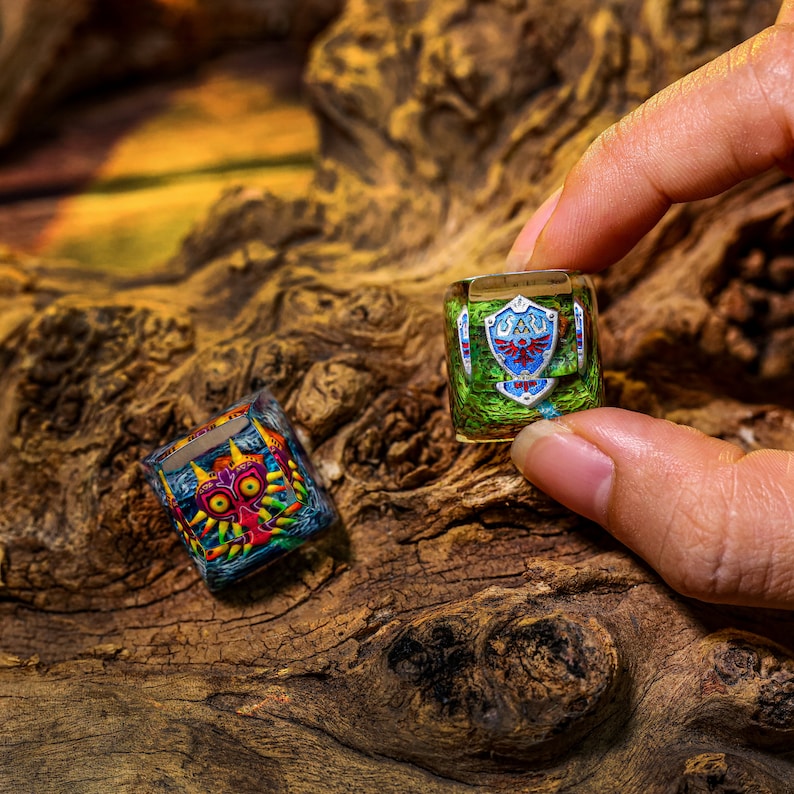 May include: Two resin keycaps featuring characters from the Legend of Zelda video game series. One keycap is green and blue with the Triforce symbol and the other is multi-colored with a red and yellow mask.