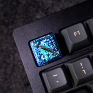 May include: A blue and green resin keycap with a stylized sword design. The keycap is on a black keyboard.