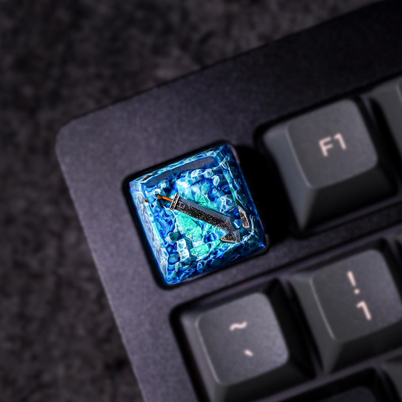 May include: A blue and green resin keycap with a stylized sword design. The keycap is on a black keyboard.