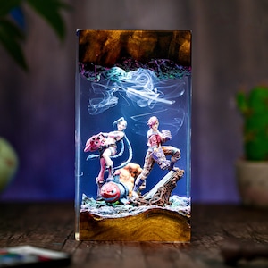 May include: A rectangular resin art piece featuring two figurines in a dynamic pose, with a pumpkin-like figure at the base. The figures are set against a backdrop of swirling white smoke and a dark, textured background.
