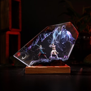 May include: A resin sculpture featuring two figures with glowing lightsabers engaged in a duel. The figures are set within a clear, angled resin block with swirling purple and blue accents, mounted on a wooden base. The overall theme is science fiction.