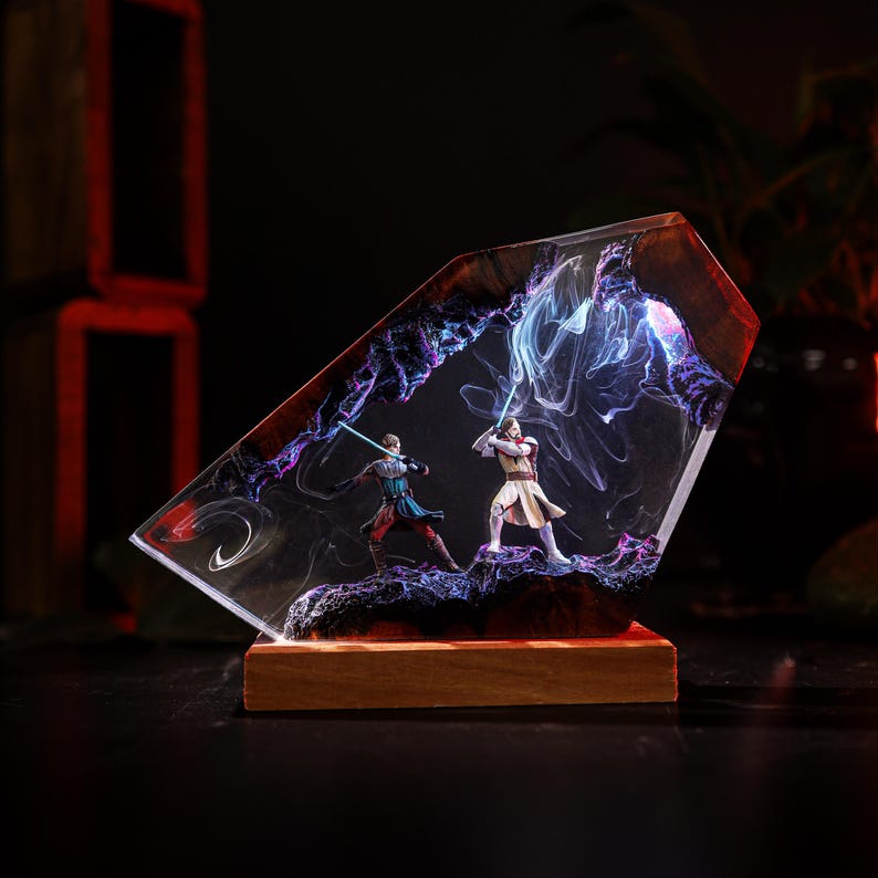 May include: A resin sculpture featuring two figures with glowing lightsabers engaged in a duel. The figures are set within a clear, angled resin block with swirling purple and blue accents, mounted on a wooden base. The overall theme is science fiction.