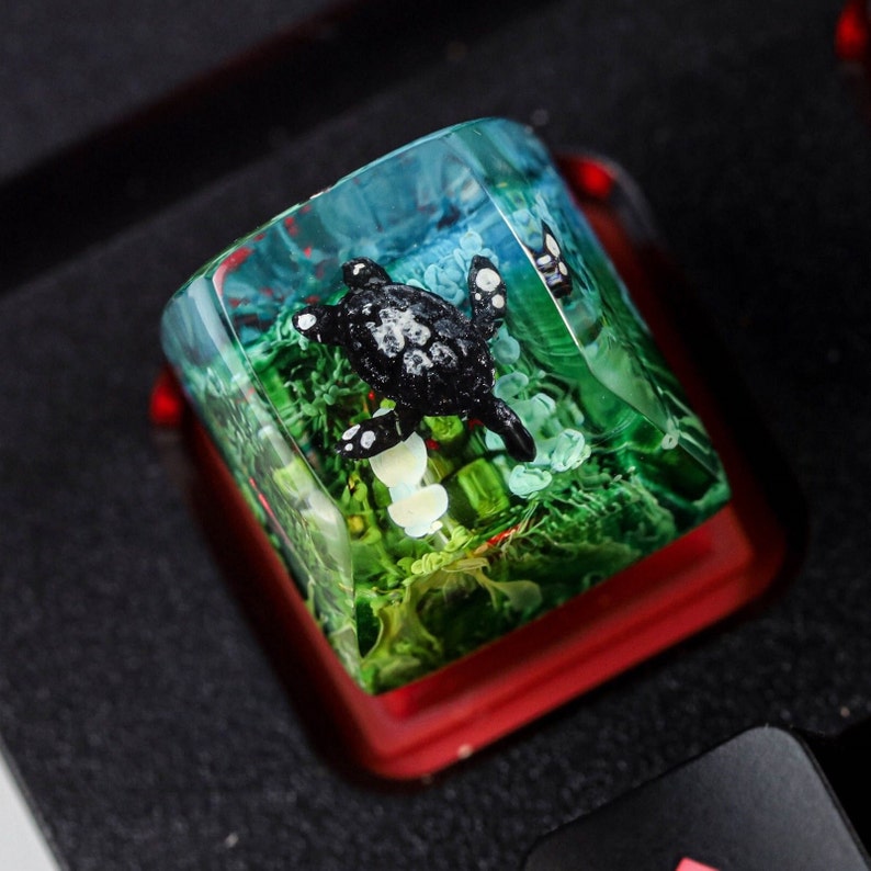 May include: A clear resin keycap with a black turtle figurine inside. The turtle is surrounded by green and blue resin, resembling an underwater scene.