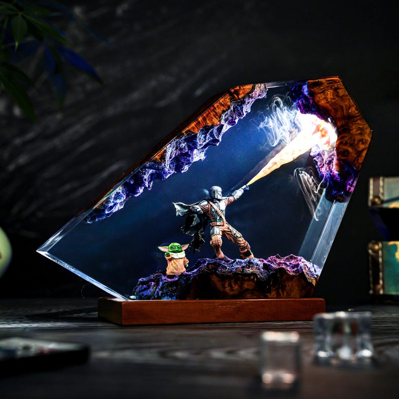 May include: A decorative resin and wood lamp featuring a miniature armored figure in a cape firing a bright orange flame, with a small green creature standing nearby. The scene is encased in a translucent, angular resin block with purple accents.