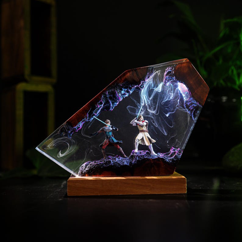 May include: A resin art piece featuring two figures with lightsabers in a dynamic pose, set within a clear, angled structure. The piece has a wooden base and a swirling, colorful background with purple and blue hues.