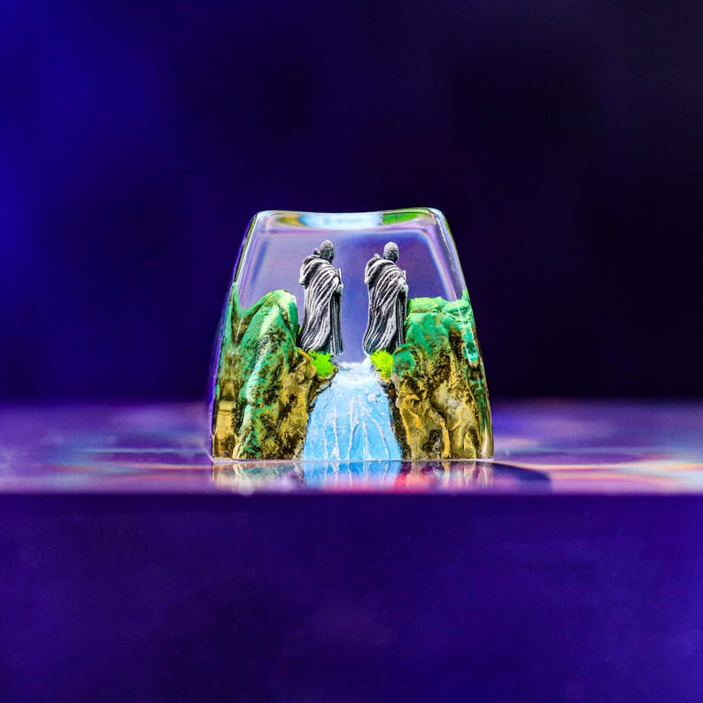 May include: A clear resin keycap with a miniature scene of two figures in cloaks standing on a green and brown landscape with a waterfall.