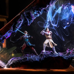 May include: A resin art piece featuring two figures with blue lightsabers engaged in a duel. The figures are set against a backdrop of swirling blue, purple, and black colors, with a dark base.