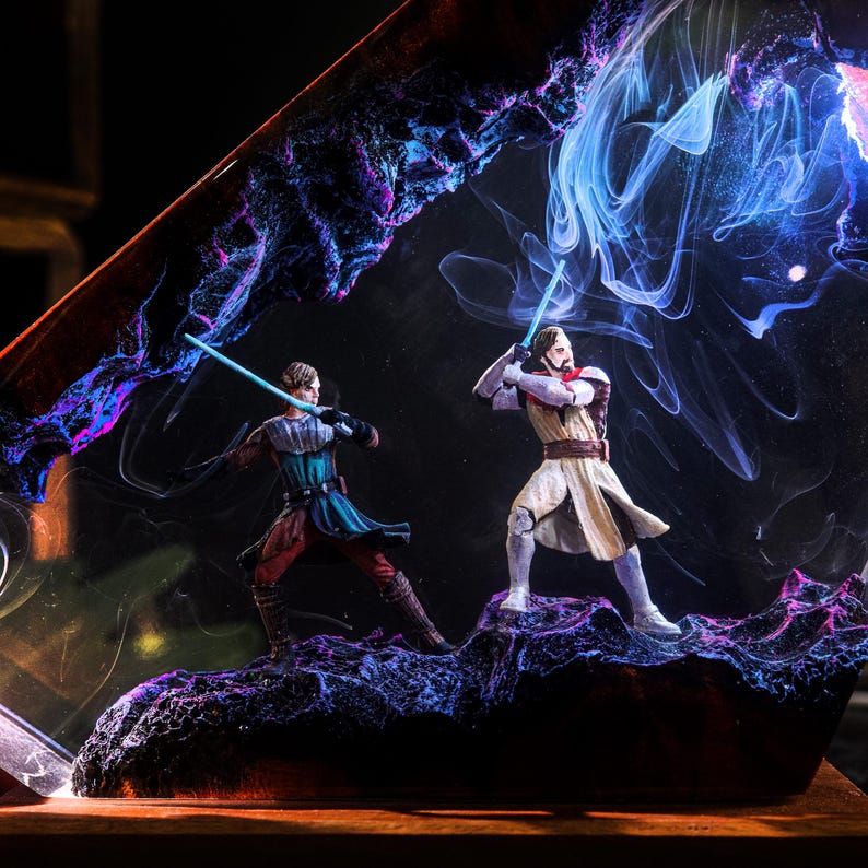 May include: A resin art piece featuring two figures with blue lightsabers engaged in a duel. The figures are set against a backdrop of swirling blue, purple, and black colors, with a dark base.